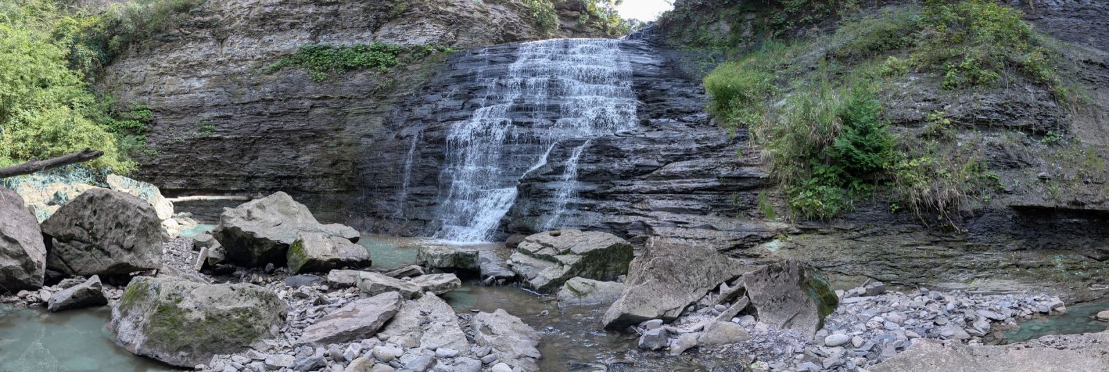 A tiered waterfall spilling down layered grey shale into a shallow pool, framed by large boulders and a steep wooded gorge in Niagara.