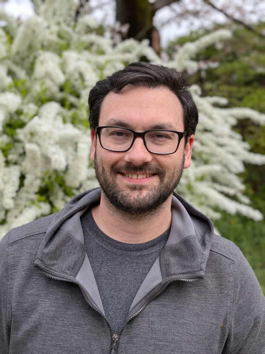 Portrait of Evan Verworn smiling at the camera in glasses and a gray jacket, with flowering trees behind him.